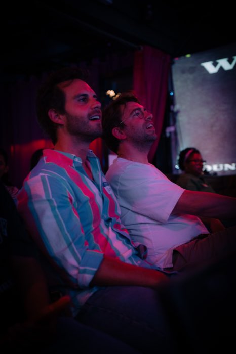 Two friends in the audience, lit pink, watching the show